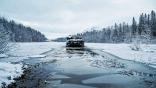 a tank pictured from behind rolling through a semi-frozen tree-lined body of water